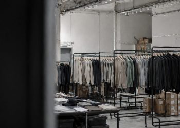 Industrial clothing storage: rows of jackets on black metal racks in a warehouse, with stacked boxes on the floor and tables of folded garments nearby.